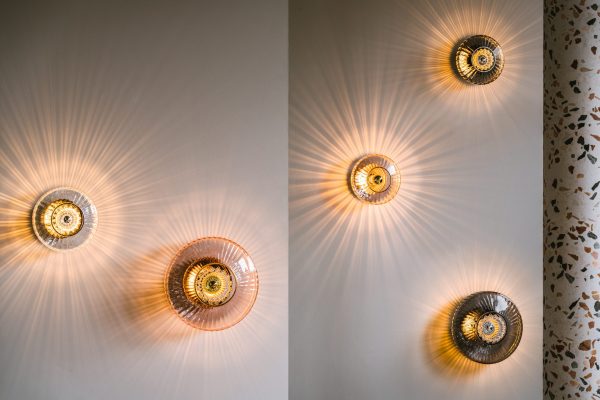 Fire DESIGN BY US - New Wave Optic Væglampe XL væglamper, der kaster strålende lysmønstre på en beige væg, hver med en smule forskellig farvetone, der spænder fra kølige til varme toner.