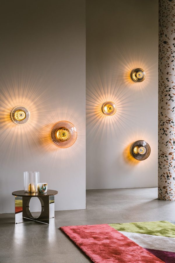 Et moderne stuehjørne med DESIGN BY US - New Wave Optic Væglampe XL, der støber varme lysmønstre på en grå væg. et lille, flerfarvet sidebord rummer dekorative genstande, suppleret med et farverigt tæppe og mønstret gardin.