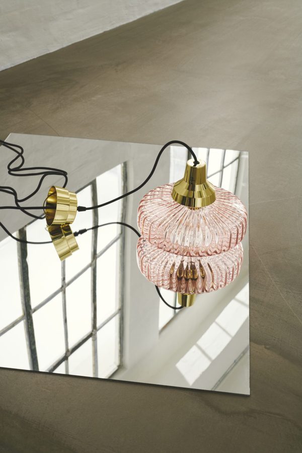 En kunstnerisk udstilling af to DESIGN BY US - New Wave Pendel-lys med guldarmaturer og indviklede lyserøde glasskærme, hængende over et spejl på et betongulv i et rum med store vinduer.