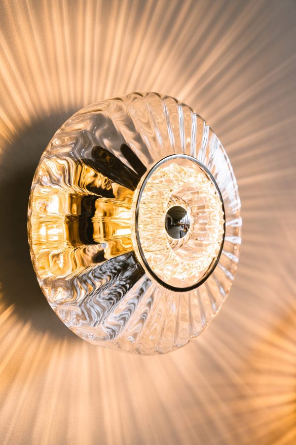 En gennemsigtig og tekstureret New Wave Optic Væglampe er oplyst og kaster et varmt, glødende lys på den beige væg bagved. Lampen udsender et mønster af lys og skygge, hvilket understreger dens indviklede design.