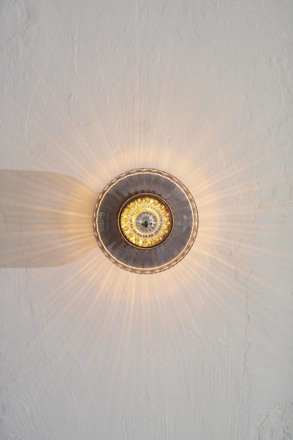 En gylden cirkulær loftslampe fra TomorrowsDesign, der støber et strålende solstrålemønster på en tekstureret hvid væg.