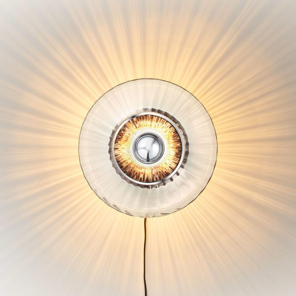 Et cirkulært DESIGN BY US - New Wave Optic Væglampe med et skarpt, centralt lys omgivet af en gennemskinnelig, tekstureret glasskærm, der kaster strålende lysstråler hen over en lys baggrund.