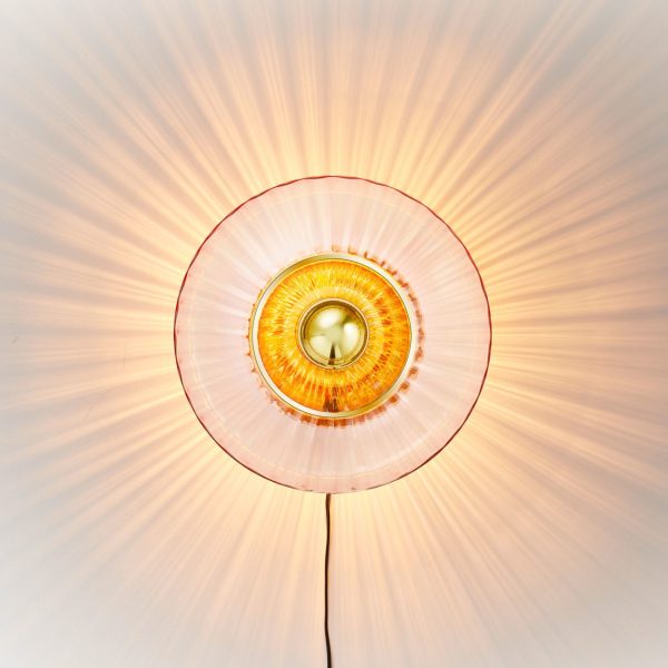 Et set ovenfra af en tændt loftslampe med et designmøbler gyldent center og udstrålende orange lysmønstre på en hvid baggrund.