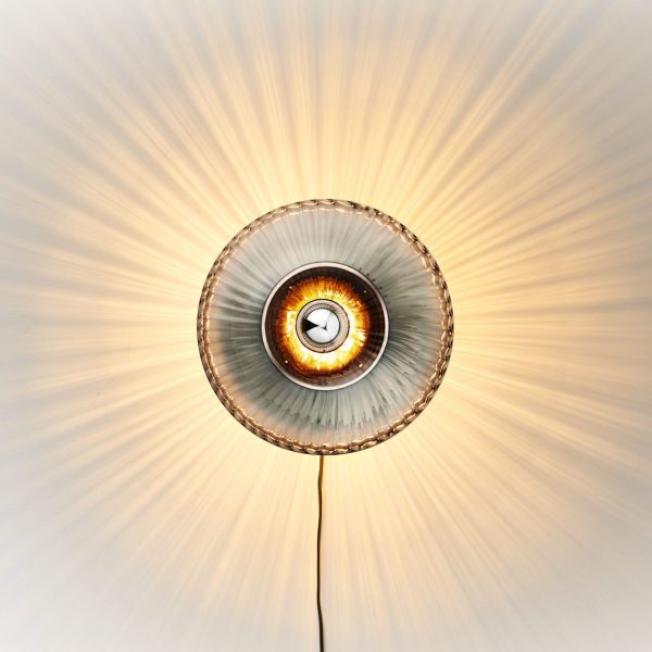 A DESIGN BY US - New Wave Optic Væglampe XL med et cirkulært, rillet glasdæksel og en glødende pære i midten, der kaster strålende lysstråler hen over loftet.