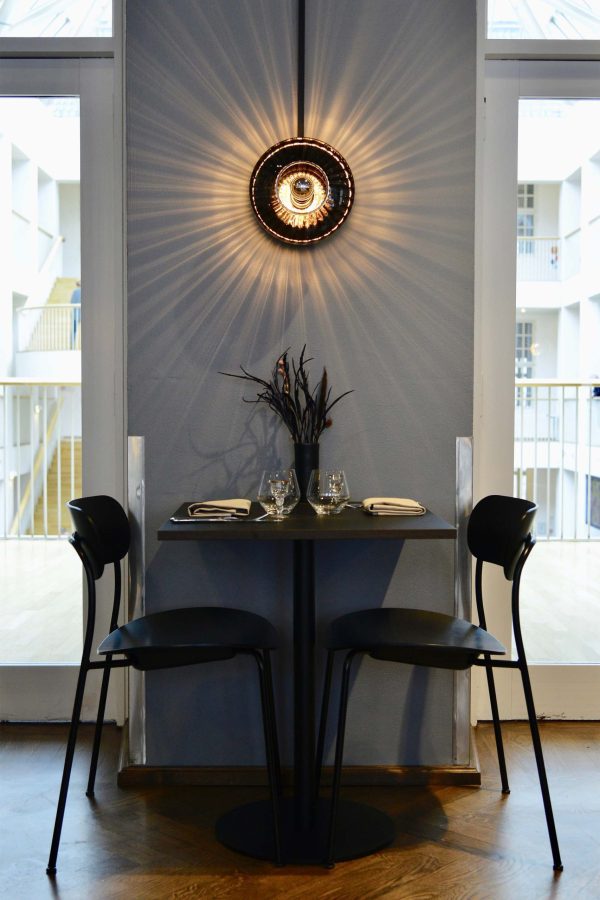 En lille spiseplads med et rundt bord med to sorte stole, designet med omhyggelig sans for detaljer, sat op mod en slående væg oplyst af et centralt cirkulært lys, der skaber en halo-effekt. Scenen er indrammet af et luftigt, moderne interiør.