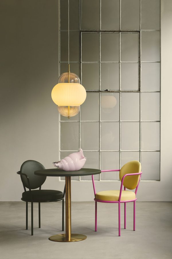 Et moderne interiør med et mørkt rundt bord, en pink griseformet skulptur, en neon pink stol og en sort stol mod en væg med firkantede spejle. ET DESIGN AF OS - Shahin XL Pendel tilføjer en blød glød.