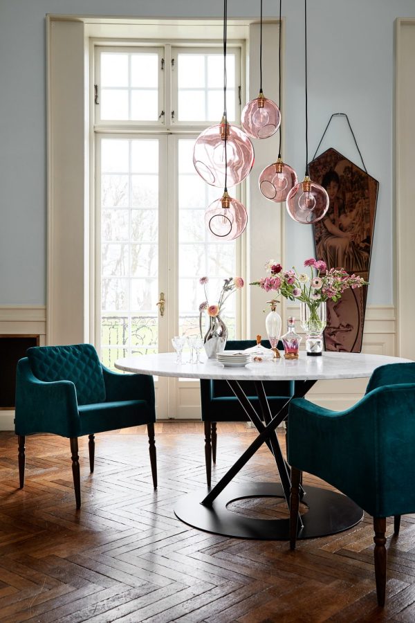 En stilfuld spisestue med blågrønne lænestole, et sort rundt bord og en række pink glaslamper. Et indrammet maleri og friske blomster tilføjer elegance, sat mod store vinduer og trægulve. Interiørdesignet forbedrer den overordnede stemning i rummet.