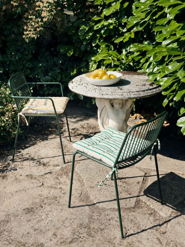 En solbelyst have med et rundt bord i sten understøttet af en klassisk skulptur som base, omgivet af to metalstole med stribede hynder. Bordet rummer en skål med gule frugter. Frodigt grønt løv opsluger baggrunden. Dette setup viser udsøgte designmøbler i udendørs omgivelser.