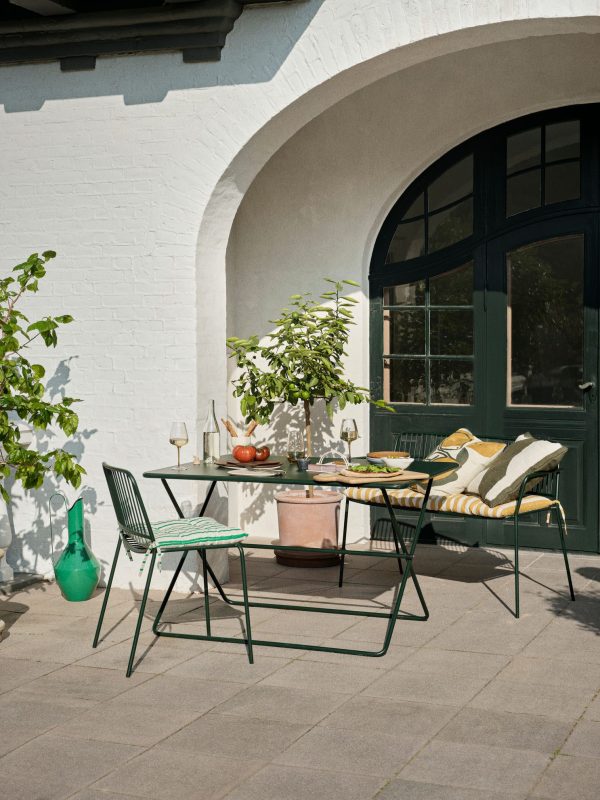 Et udendørs terrasseområde med et designbord i metal med en grøn stol og en lænestol med stribede hynder, placeret ved siden af potteplanter og under buegange. En klar dag med solskin forbedrer scenen.
