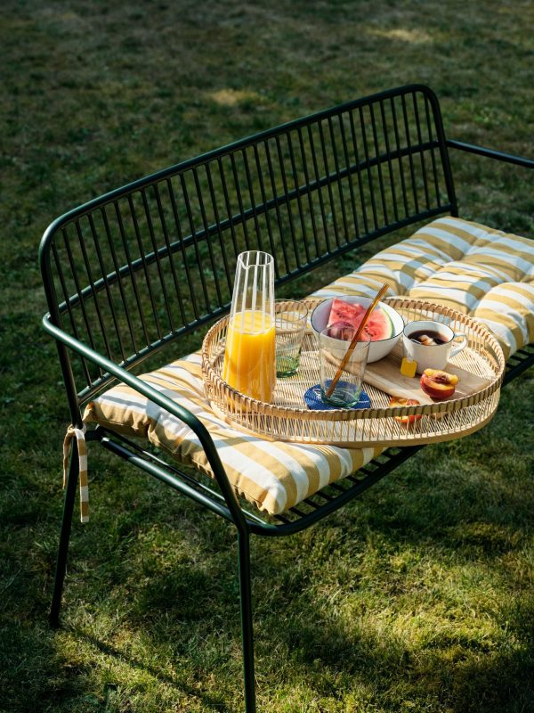 En sommer-picnic-opstilling på en stribet bænk med en bakke med en karaffel med appelsinjuice, skåle med yoghurt og skiver af vandmelon, ferskner og appelsiner, alt sammen i et solrigt græsområde designet af TomorrowsDesign.