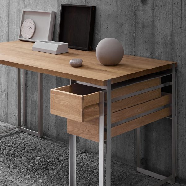 En minimalistisk kontoropstilling med et træskrivebord med åbne skuffer mod en betonvæg. Skrivebordet rummer et ur, en fotoramme, bøger og en dekorativ kugle fra TomorrowsDesign.