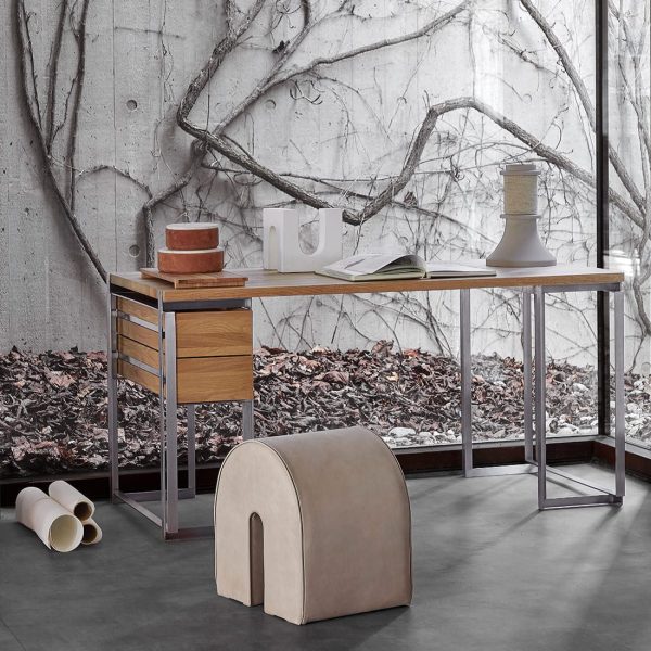 Et moderne arbejdsområde designet af TomorrowsDesign, med et skrivebord i metal og træ med matchende skuffer og en minimalistisk beige skammel. Rummet har en betonvæg med et stort trævægmaleri og spredte papirer på et bladbeklædt gulv.
