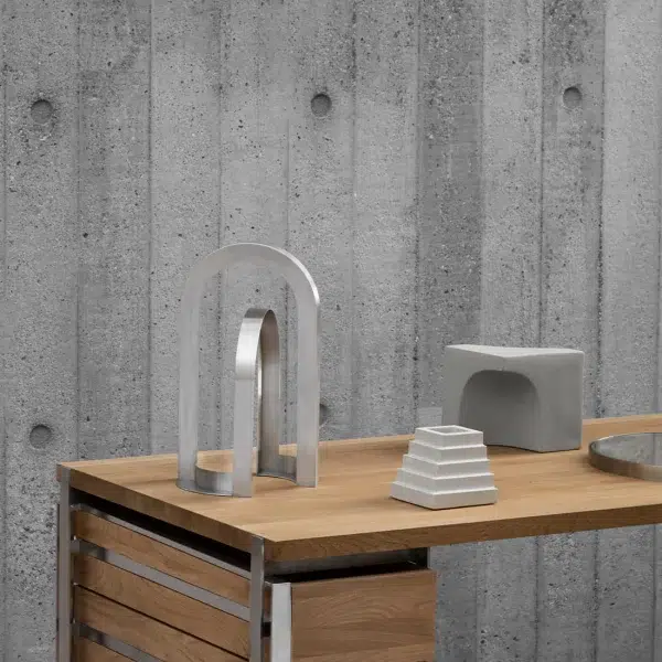 Et minimalistisk arrangement på et træbord med en gennemsigtig buet skulptur, en grå blok og en lille hvid trappepyramide, mod en betonvægbaggrund viser udsøgt designmøbler-æstetik.