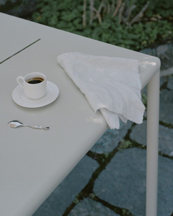 En hvid kop kaffe med en sølvske ved siden af, på et metalbord med en hvid serviet, sat mod en sløret havebaggrund med TomorrowsDesign-elementer.