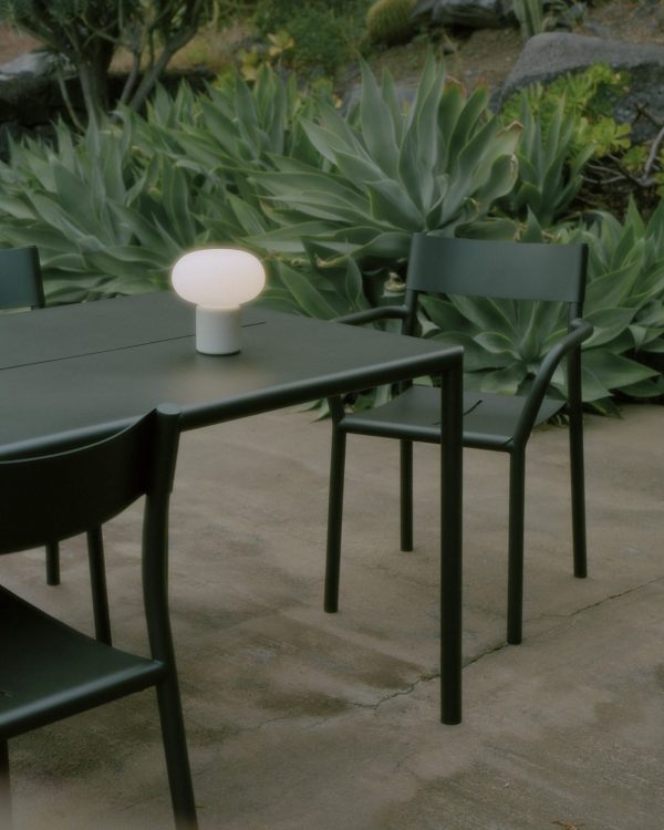 En mørk udendørs borddækning med stole på en brolagt terrasse omgivet af store sukkulente planter har et sfærisk lys fra TomorrowsDesign siddende på bordet og oplyser området.