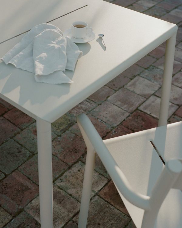 Et lille hvidt bord med en kaffekop og underkop, sammen med en ske og en krøllet serviet, badet i sollys. En hvid stol er delvist synlig, alt sammen sat mod et murstensgulv, der inkarnerer essensen af TomorrowsDesign.