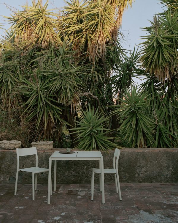 Et lille udendørs opholdsområde med hvid designmøbler inklusiv bord og to stole, sat på baggrund af frodige grønne palmelignende planter under en klar himmel. Indstillingen fremstår rolige og naturlige.
