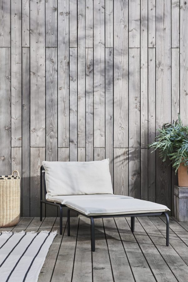 Et minimalistisk udendørs siddeområde med en OYOY - Furi Udendørs Ottoman med en hvid pude, sat op mod en forvitret træplankevæg. et stribet tæppe og en plante i en kurv forstærker den hyggelige stemning.