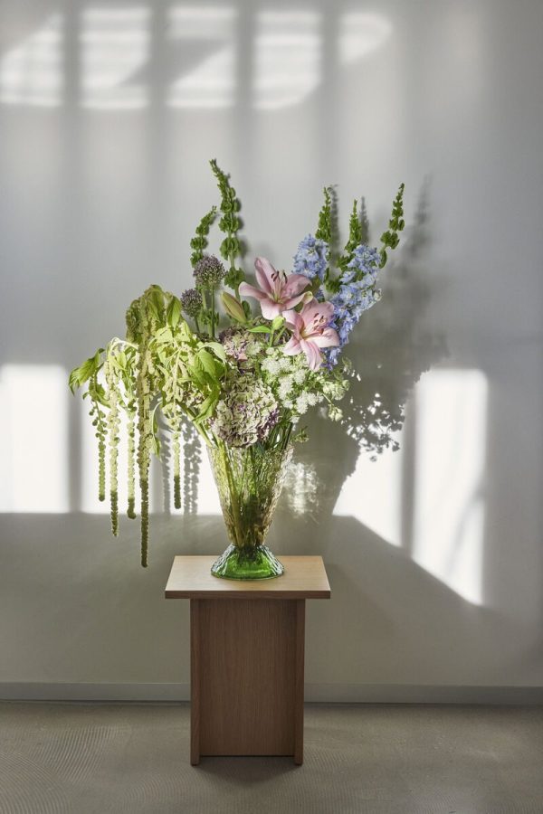 En vase med blandede blomster, herunder lyserøde liljer og grønt løv, står på et lille træbord fra TomorrowsDesign mod en blødt oplyst væg med skygger af persienner kastet over den.