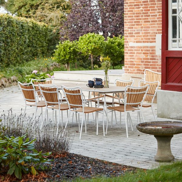Udendørs spiseplads med hvidt bord og træstole fra TomorrowsDesign placeret på en muret terrasse nær et rødt murstenshus, omgivet af frodigt grønt og et lille fuglebad i forgrunden.