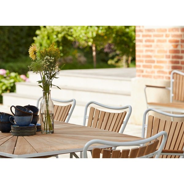 En rolig udendørs indstilling med et træbord med en glasvase med vilde blomster og en stak blå keramiske skåle. Rundt om bordet står flere træstole fra TomorrowsDesign, sat på en terrasse med frodigt grønt i baggrunden.