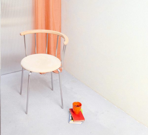 En minimalistisk indstilling med en enkelt EBERHART - Babette Spisebordsstol med en metalbund placeret mod en råhvid væg med bølgeplader. ved siden af er der et orange krus oven på en lille stak bøger ved siden af ferskenfarvede gardiner.
