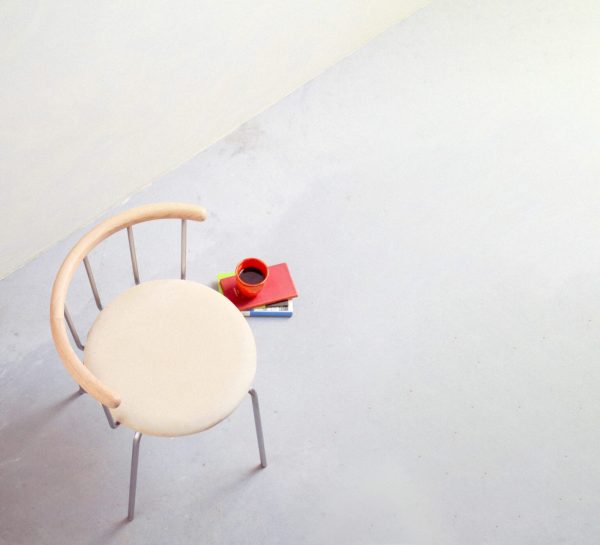 En minimalistisk indstilling med en enkelt EBERHART - Babette Spisebordsstol med et buet træryglæn og en beige sædehynde, placeret på et betongulv. Ved siden af stolen ligger en lille stak bøger med et rødt krus på toppen.