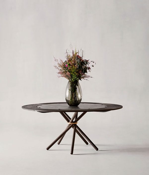 En minimalistisk indstilling med et rundt, mørkt træ EBERHART - Hector 140 Tillægsplader bord med en vase med vilde blomster centreret på toppen, placeret mod en almindelig, lysegrå baggrund.