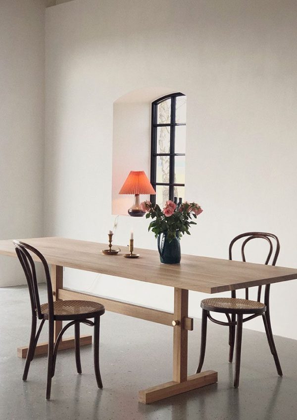 En minimalistisk spiseplads med en EBERHART - Gaspard Spisebord 240, to stokstole, et højt sortrammet spejl lænet op ad væggen og en vase med blomster under en blød pink lampeskærm.