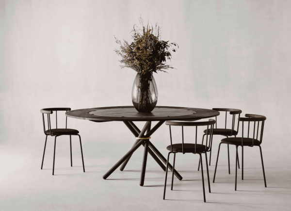 En minimalistisk spisestue med et rundt, mørkt træbord med en unik metalbund, omgivet af fire EBERHART - Babette Spisebordsstol stole. en stor vase med tørrede buskede blomster står oven på bordet, sat mod en almindelig, lys baggrund.
