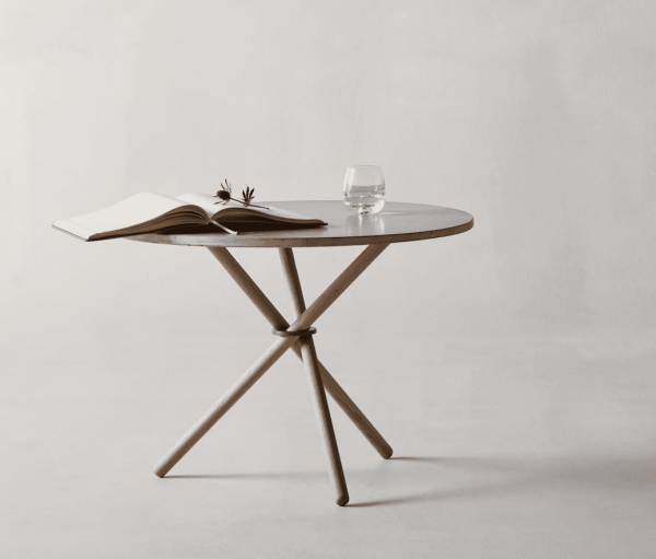 Et EBERHART - Daphne Sofabord Ø65 og et par runde sorte briller hviler på et lille, rundt bord med et krydsende metalbendesign, ved siden af et klart glas vand, mod en lysegrå baggrund.