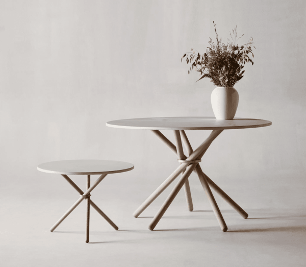To runde, minimalistiske møblerborde med krydsede træben, et mindre og et større. Det større bord rummer en hvid vase med tørrede planter. Baggrunden er en almindelig, lys væg.