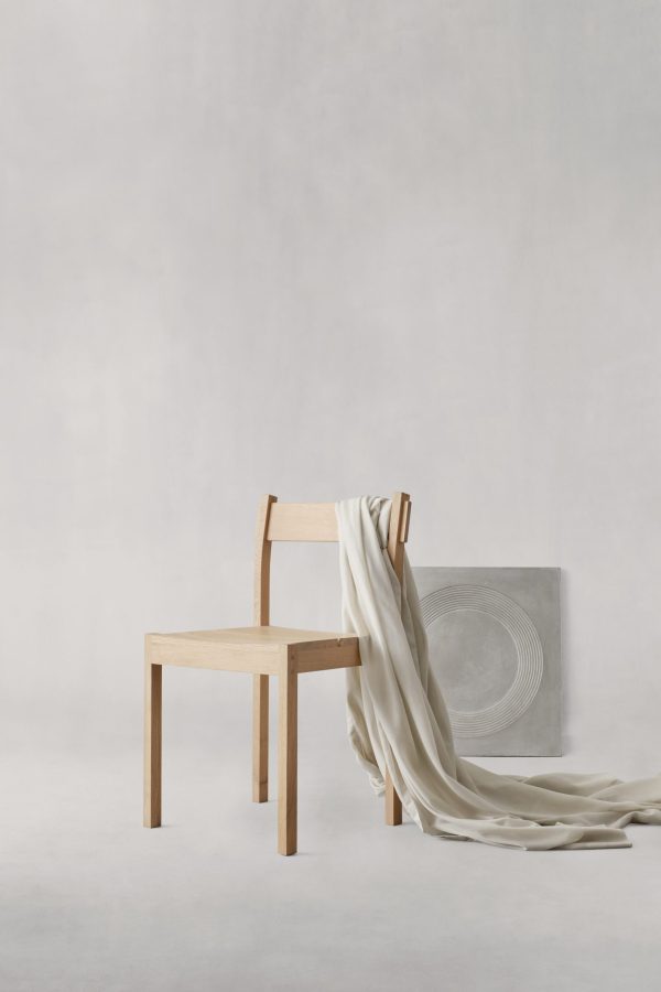 Et minimalistisk setup med en EBERHART - Thibault Spisebordsstol med et cremet stof draperet over den ene side. Ved siden af stolen er der en grå cylindrisk skulptur med et cirkulært reliefmønster, alt sammen mod en lysegrå baggrund.