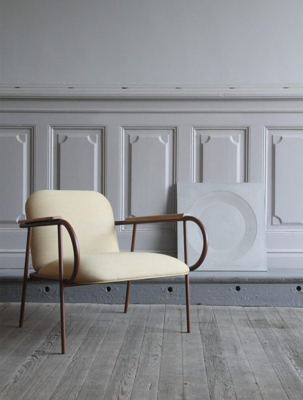 En moderne lænestol med beige polstring og træarme står på et gråt trægulv mod en hvid panelvæg i et værelse med et enkelt, elegant design. EBERHART - Fausto Lænestol stolen er den perfekte tilføjelse til dette rum.