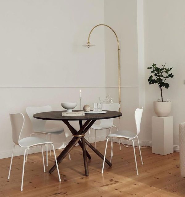 En minimalistisk spisestue med et rundt mørkt bord, fire hvide designmøbler stole og en gylden buet gulvlampe. En potteplante er i hjørnet og en simpel pendel hænger ovenover.