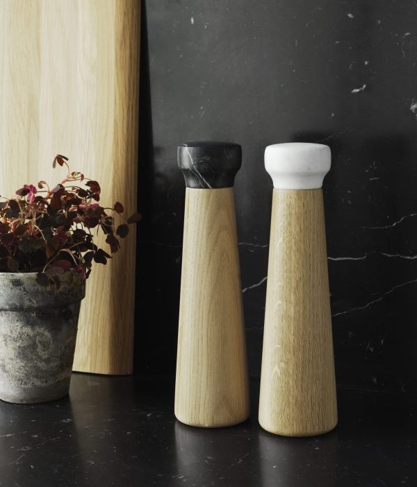 To NORMANN COPENHAGEN Craft Peberkværn salt- og peberkværne, en med sort top og en med hvid top, står på en sort marmorflade ved siden af en potteplante med rødlige blade.