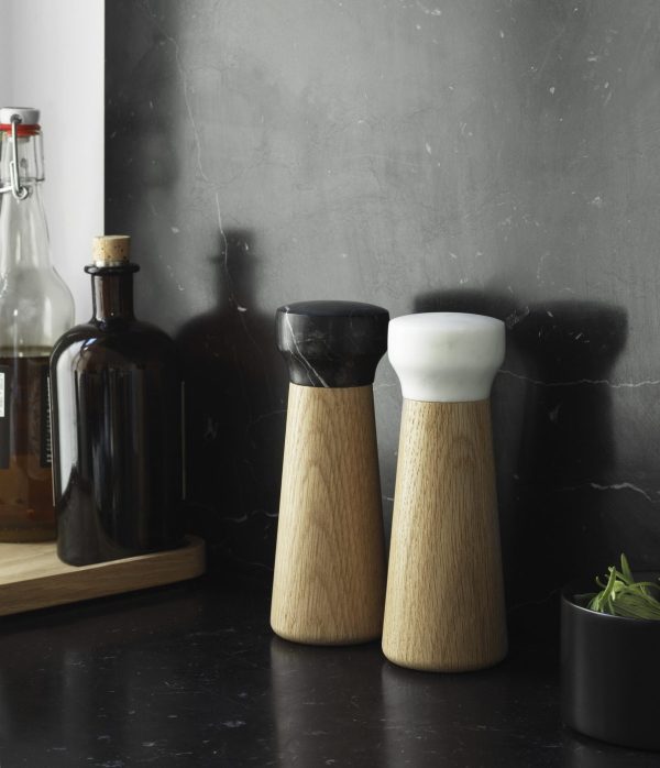 To NORMANN COPENHAGEN - Craft Peberkværn med sorte og hvide toppe er placeret mod en mørkegrå tekstureret baggrund, ledsaget af en brun flaske og en lille sort skål med urter.