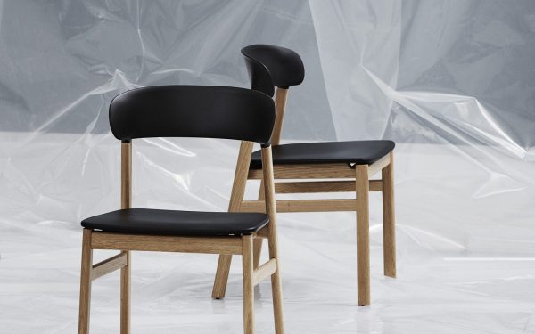 To NORMANN COPENHAGEN - Herit Spisebordsstol stole med træstel og sort polstring, placeret på et blankt gulv mod en draperet plastbaggrund.