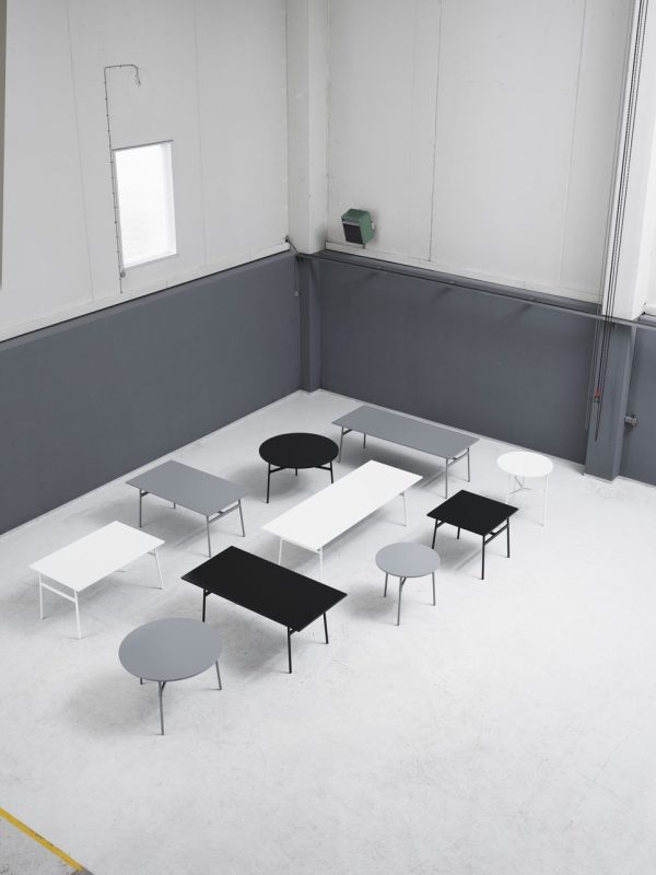 Værelse i minimalistisk stil med et arrangement af flere hvide og sorte rektangulære borde med runde skamler, placeret på et gråt gulv med en gul streg. Rummet har hvide vægge, høje vinduer og fremviser elegante designmøbler.