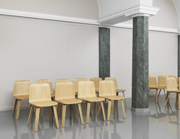 En række enkle træstole arrangeret pænt op ad en væg i et rummeligt rum med blanke hvide gulve, suppleret med elegante grønne marmorsøjler og et dekorativt hvidt loft, der viser gennemtænkte designelementer.