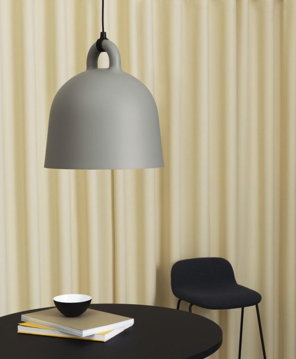 Et moderne interiør med en stor NORMANN COPENHAGEN - Bell Lampe Ø55 hængende over et sort rundt bord med en sort stol, en bog og en skål på bordet, på baggrund af cremefarvede gardiner.