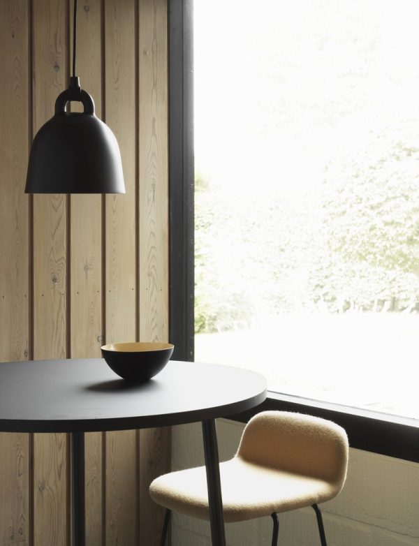 Et minimalistisk interiør med en rund sort NORMANN COPENHAGEN - Bell Lampe Ø22 med en sort skål på, en beige tekstureret stol og en stor sort pendel, ved siden af et stort vindue med udsigt til et grønt ydre.