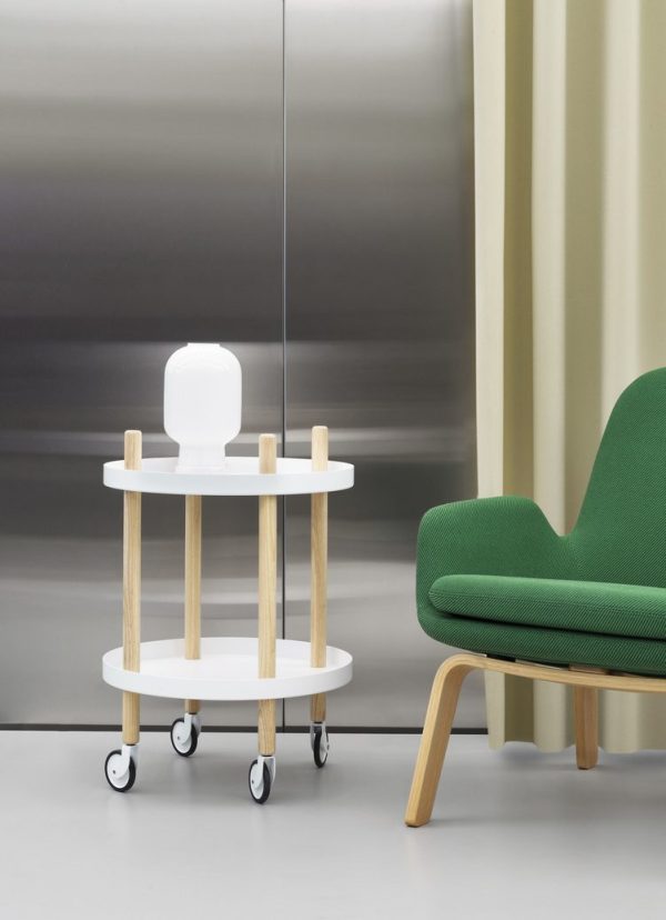 Et moderne interiør med en hvid cirkulær vogn med tre træstænger og små sorte hjul, ved siden af en elegant grøn lænestol designet med møbler-æstetik, på baggrund af stålgrå og cremefarvede gardiner.