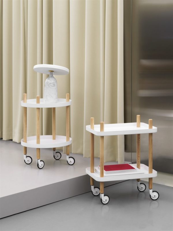 To moderne runde rullende rullevogne med hvide lag med træstandere, den ene med bøger og en hvid vase, placeret foran draperede beige gardiner og en sløret baggrund, perfekt tilføjelse til ethvert interiør.