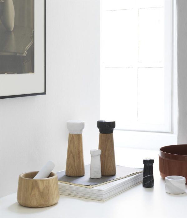 Et moderne køkkenhjørne med naturligt lys, der strømmer ind fra et vindue, med en samling af stilfuldt køkkenudstyr, herunder salt- og peberkværne i træ, en morter og støder og NORMANN COPENHAGEN Craft Peberbøsse på en bog.
