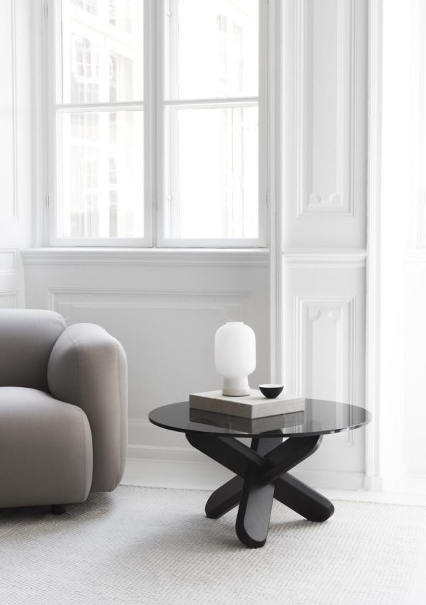 En minimalistisk stue med møbler som en beige sofa, et sort rundt sofabord, et hvidt skulpturelt stykke og en kop. Store hvidrammede vinduer giver udsigt til bygninger udenfor og skaber et lyst og luftigt rum.
