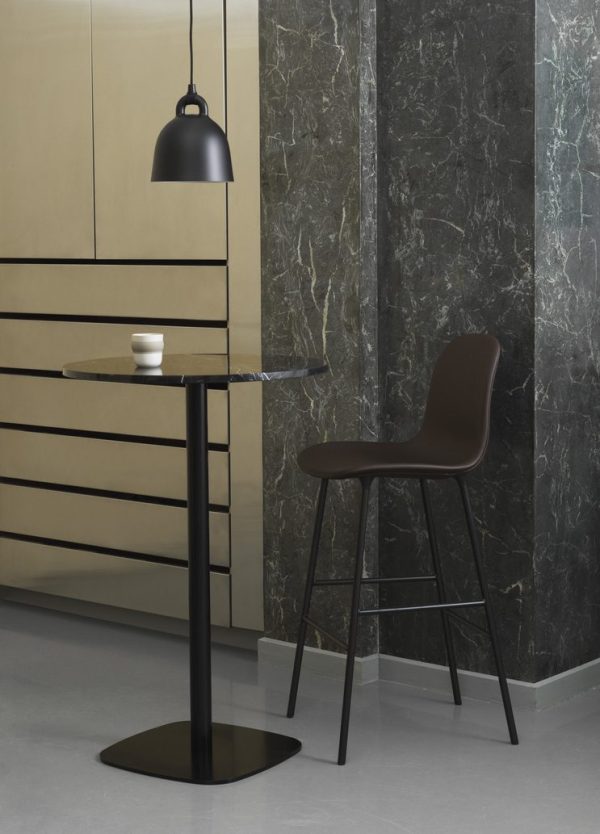 Et moderne køkkenhjørne med en mørkebrun barstol ved et sort højbord nær et beige skab og en mørk marmorvæg. En hængende NORMANN COPENHAGEN - Bell Lampe Ø22 oplyser scenen, med to hvide kopper på bordet.