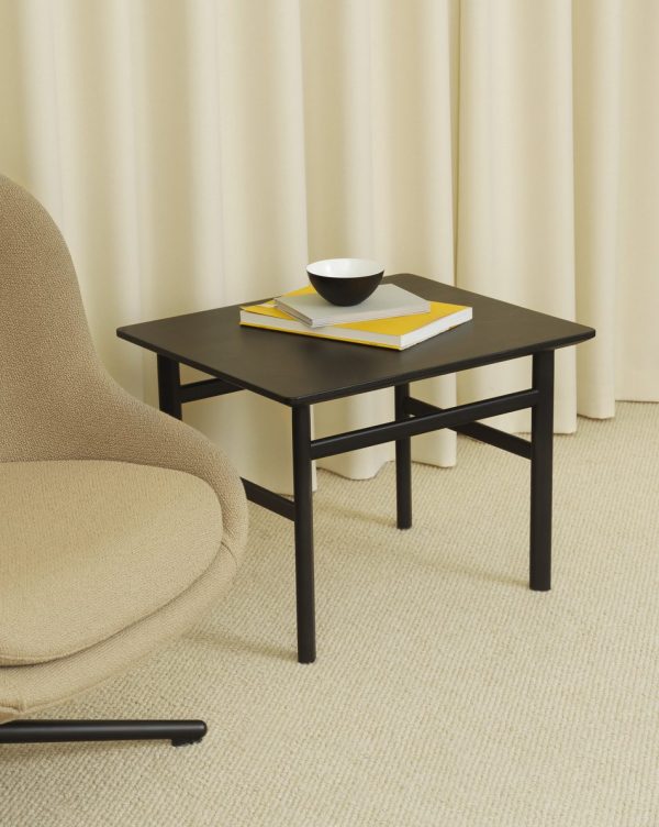 Et lille sort bord med en skål og to bøger stablet på, placeret ved siden af en beige lænestol og mod en baggrund af cremefarvede gardiner. Gulvet er belagt med et lyst beige tæppe, der forstærker rummets interiørdesign.