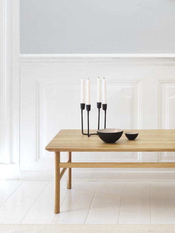 Et minimalistisk interiør med en bænk af naturligt træ mod en hvid panelvæg med et trægulv. På bænken er der en sort lysestage med tre hvide lys og to sorte møbler skåle.