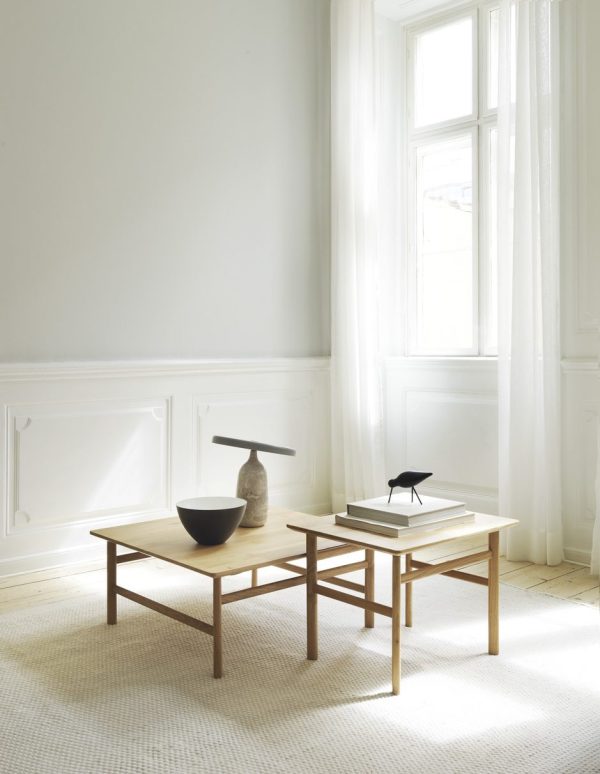 Et minimalistisk interiør med to lyse træborde med en grå vase, en sort fuglefigur og bøger på toppen. Rummet har hvide vægge, gulvlange gardiner fra TomorrowsDesign og et stort vindue, der slipper naturligt lys ind.